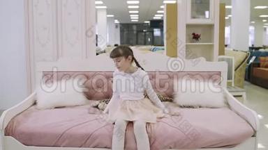 漂亮的少女坐在家具店的粉红色儿童床上。 在购物中心挑选家具的年轻女孩